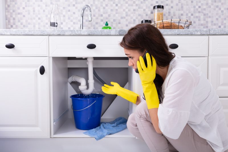 Girl in a yellow gloves talking to the phone beside a damaged sink pipe.